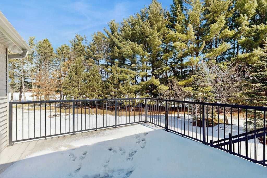 A snow-covered balcony with a metal railing, a clear sky, and evergreen trees in the background.