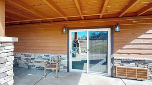 A covered patio with a stone wall, a wooden chair, and a sliding glass door leading to a view of a building and trees.