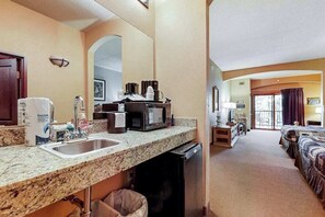 A hotel room with a kitchenette, a sink, a microwave, a coffee maker, a trash can, a bed, a desk, and a window with curtains.