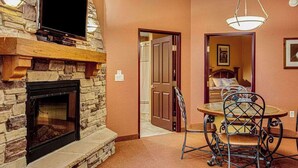 Una habitación con chimenea de piedra, un televisor en la pared, una mesa de comedor con sillas y una puerta que da a otra habitación.
