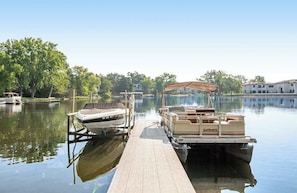A dock with a boat and a pontoon boat, surrounded by calm water and trees. - Lakefront condo with beach access, firepit, games, fireplace, & dock (Wisconsin Dells)