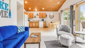 Ein modernes Wohnzimmer mit einem blauen Sofa, einem grauen Sessel und einem Couchtisch aus Holz. Im Hintergrund ist eine Küche mit Holzausstattung und ein Essbereich mit Tisch und Stühlen zu sehen.