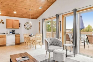 A modern interior with a wooden ceiling, a dining area with wicker chairs, a grey armchair, and a balcony with outdoor furniture.
