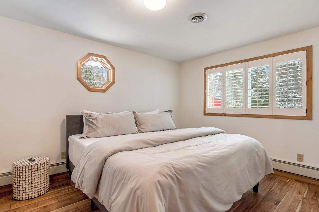 Un dormitorio con una cama, un espejo redondo, una ventana con persianas y un taburete pequeño y redondo.