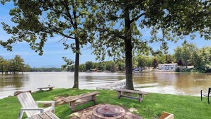 A lakeside outdoor seating area with a fire pit, benches, and a view of a lake and distant buildings. - Lakefront retreat with panoramic views, large backyard, & direct beach access (Wisconsin Dells)