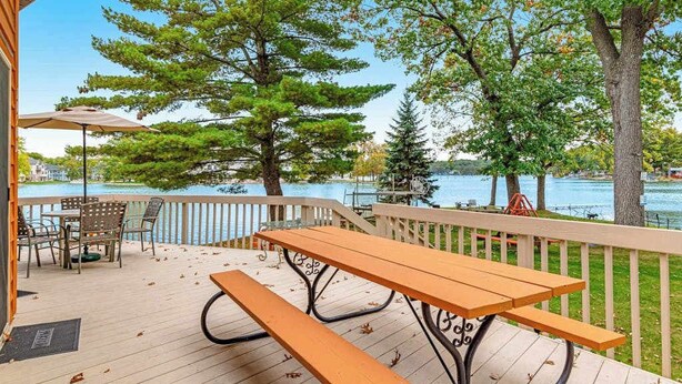 A wooden deck with picnic tables and chairs, overlooking a body of water with trees. - Lakefront retreat with panoramic views, large backyard, & direct beach access (Wisconsin Dells)