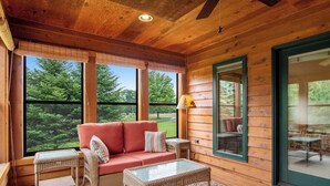A wooden cabin interior with a sofa, wicker furniture, and large windows offering a view of greenery. - Dog-friendly lakefront getaway with dock, firepit, wet bar, game room, & AC (Eagle River)