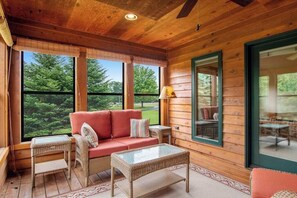 Ein Holzblockbau-Innenraum mit einer Couch, Korbmöbeln und großen Fenstern mit Blick auf Grünflächen.