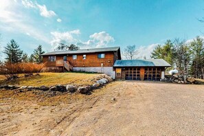 A two-story log cabin with a garage, surrounded by a gravel driveway and a stone border.