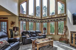 Una sala de estar acogedora con un sofá de cuero, una mesa de centro de madera y ventanas grandes con vistas a árboles.
