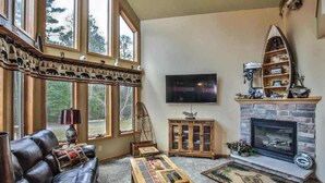 Una sala de estar con chimenea, televisor, sofá de cuero y un ventanal con vistas a árboles.