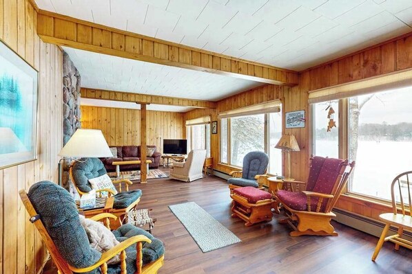 A cozy cabin interior with wooden walls, a fireplace, a television, and a large window overlooking a snowy landscape. - Lakefront Lodge w/ 2 additional cabins, private dock, firepit, kayak & hammock (Woodruff)