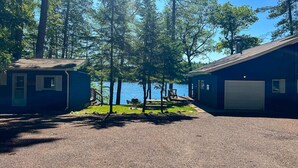 En grusvej fører til et sommerhus med garage, omgivet af træer og udsigt til en sø.