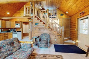 A cabin interior with a wooden staircase, a sofa, a chair, a small table, and a microwave in the kitchen area.