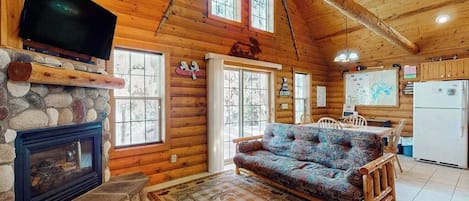 A log cabin interior with a stone fireplace, a television mounted on the wall, a patterned rug, wooden furniture, and a kitchen area.