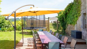 An outdoor patio with a long table and chairs, a yellow umbrella, and a stone wall with greenery. - Gîte le Caddet – Traditional Béarnais Farmhouse for Groups and Families (Poursiugues-Boucoue)