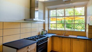 A kitchen with a gas stove, oven, wooden cabinets, a window with a view of greenery, and a pendant light. - Gîte le Caddet – Traditional Béarnais Farmhouse for Groups and Families (Poursiugues-Boucoue)
