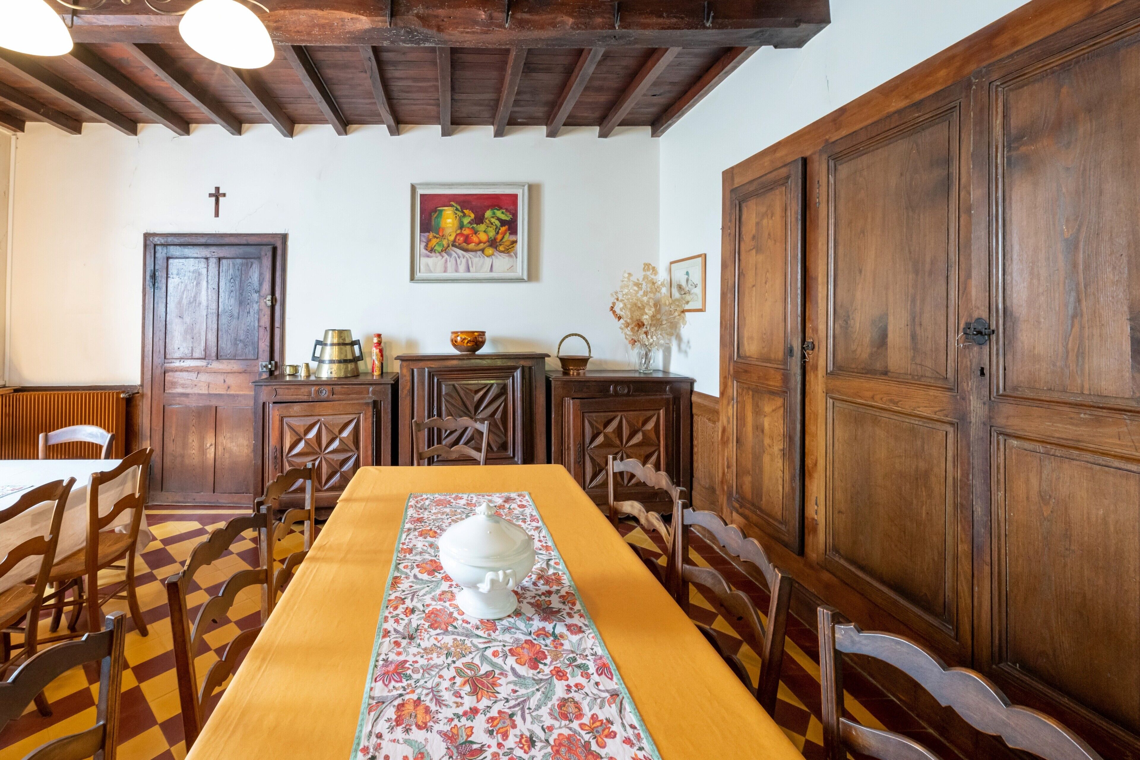 Un coin repas avec une longue table en bois, des chaises, un chemin de table à motifs floraux, des meubles en bois et une croix au mur.