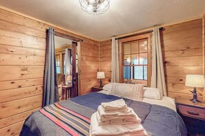 Una habitació de fusta amb un llit, dues làmpades de nit, una finestra amb cortines i una porta.