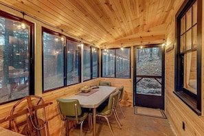 A wooden cabin interior with a dining table, chairs, and large windows offering a view of the forest.