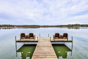 Eine Holzstele mit zwei Stühlen, die auf einen ruhigen See blicken, umgeben von Bäumen.
