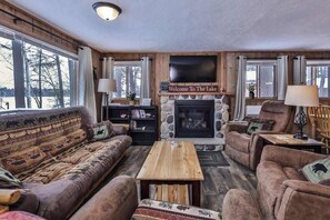 A cozy living room with a fireplace, a sofa set, a coffee table, and a television. - Snowy lake-view cabin with fireplace, full kitchen, TV, dock, & DVD player (St. Germain)