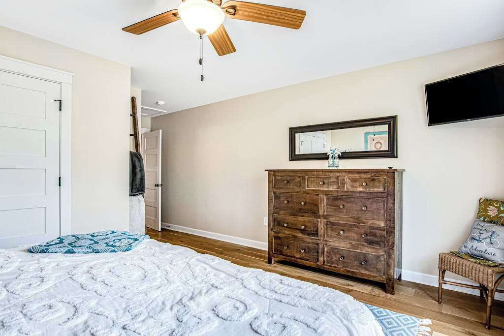 Une chambre à coucher avec un lit, une commode en bois, un ventilateur de plafond, un téléviseur et une porte.