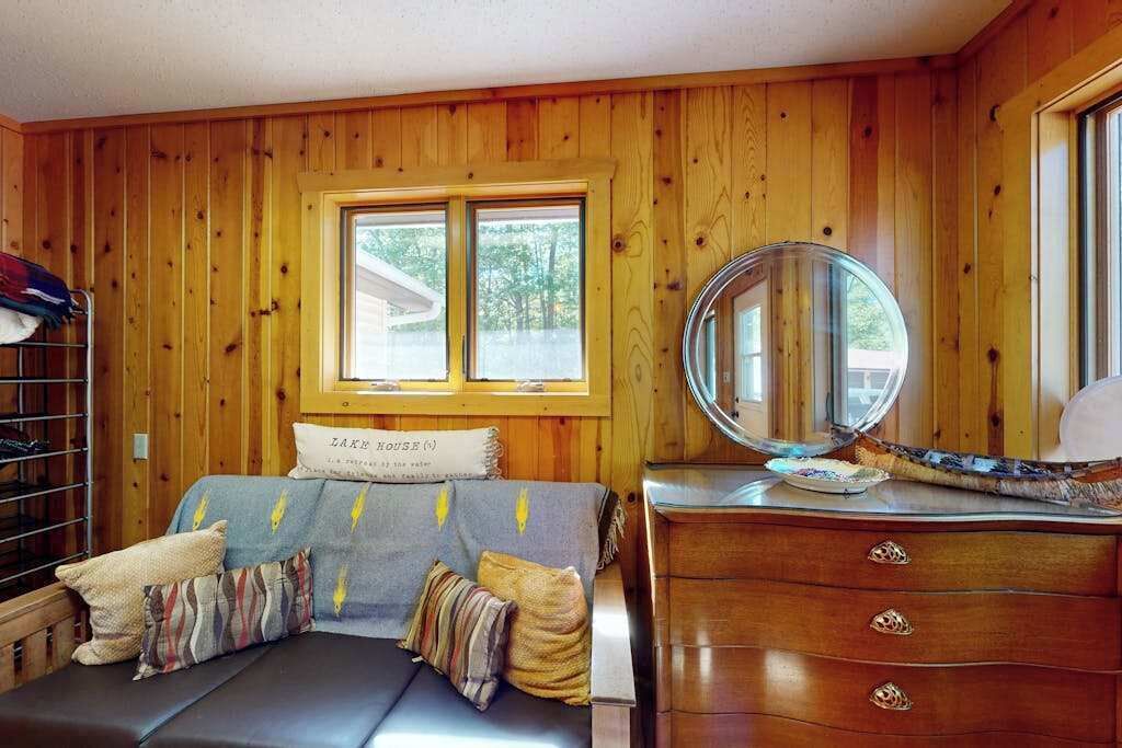 A room with wooden walls, a bed with a headboard labeled 'LAKE HOUSE,' a dresser with drawers, a mirror, and a window with a view of trees.