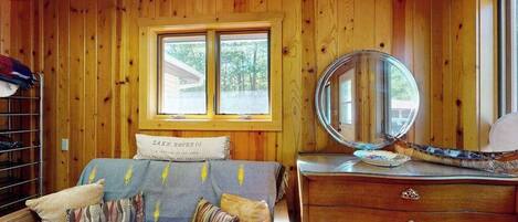 A room with wooden walls, a bed with a headboard labeled 'LAKE HOUSE,' a dresser with drawers, a mirror, and a window with a view of trees.