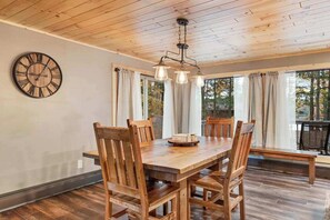 A wooden dining table with chairs, a wall clock, and a hanging light fixture in a room with wooden flooring and large windows. - Renovated lake-view retreat with full rec lake access, boat slips, & firepits (St. Germain)