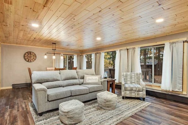 A living room with a wooden ceiling, a grey sofa, a plaid armchair, and a round ottoman. - Renovated lake-view retreat with full rec lake access, boat slips, & firepits (St. Germain)