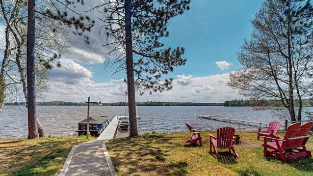 A lakeside deck with Adirondack chairs, a hammock, and a dock extending into the water. - Rustic Cozy lakefront Cabin with dock, fire pit, gas grill, W/D & quiet location (St. Germain)