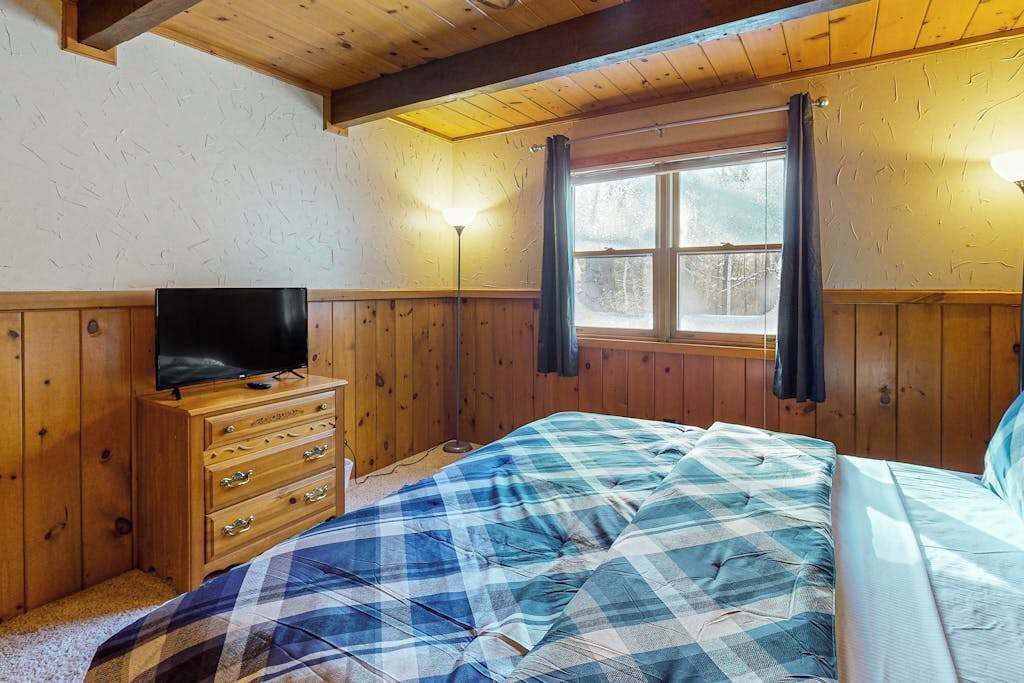 A room with wooden walls, a bed with a blue and white checkered bedspread, a wooden dresser with a television, and a window with curtains.