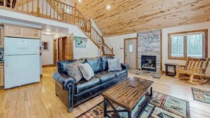 A spacious living area with a wooden ceiling, a fireplace, a black leather sofa, and a wooden rocking chair. - Rustic resort cabin with lake views, deck & access to docks, canoe, paddle boats (Mercer)