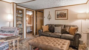 Ein gemütliches Wohnzimmer mit einem Ledersofa, einem gemusterten Sessel und einem gesteppten Ottomanen. An der Wand befindet sich ein Bücherregal mit Büchern und ein gerahmtes Bild.