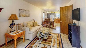 Een woonkamer met een bank, voetenbankje, salontafel, lamp, televisie en een keuken op de achtergrond.