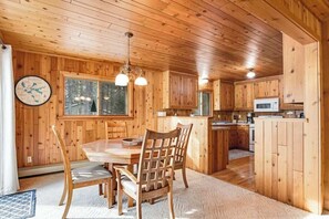 A wooden cabin interior with a dining table, chairs, a kitchen area, and a large window. - Fantastic waterfront abode with wraparound deck, dock, & WiFi - dog-friendly (Manitowish Waters)