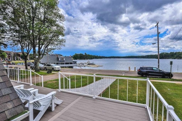 A deck with chairs overlooking a lake, a dock with boats, and a house with a grey roof.
