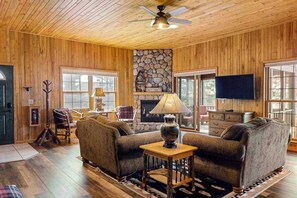 A cozy living room with wooden walls, a stone fireplace, a ceiling fan, and a television.