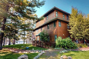 A two-story wooden cabin with a deck, surrounded by trees and a lake view.