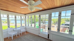 A sunroom with a ceiling fan, large windows, a white chair, a small table, and a view of a lake. - Charming lakefront home with shared dock, porch, & sunroom - walk downtown (Minocqua)