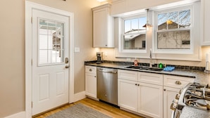 A kitchen with white cabinets, a stainless steel dishwasher, a gas stove, and a window with a view of a house. - Charming lakefront home with shared dock, porch, & sunroom - walk downtown (Minocqua)