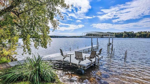 A lakeside dock with chairs and tables, surrounded by greenery and a clear sky. - Charming lakefront home with shared dock, porch, & sunroom - walk downtown (Minocqua)