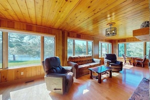 A spacious room with wooden walls and ceiling, large windows offering a view of trees, and a cozy seating area with a sofa and chairs.