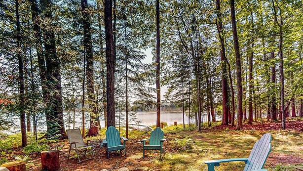 Ett skogsområde med en sjö, tre Adirondack-stolar, en lägereld och en träbänk.