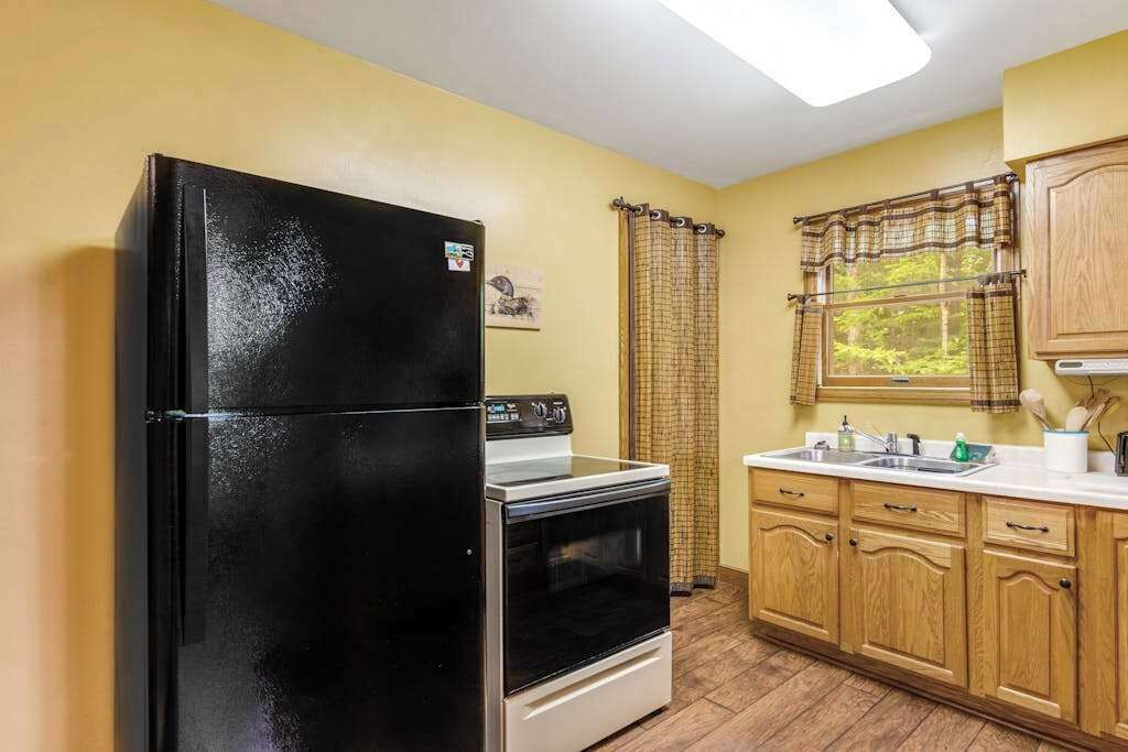 Una cocina con un refrigerador negro, un horno blanco, armarios de madera, una ventana con vistas y una cortina.