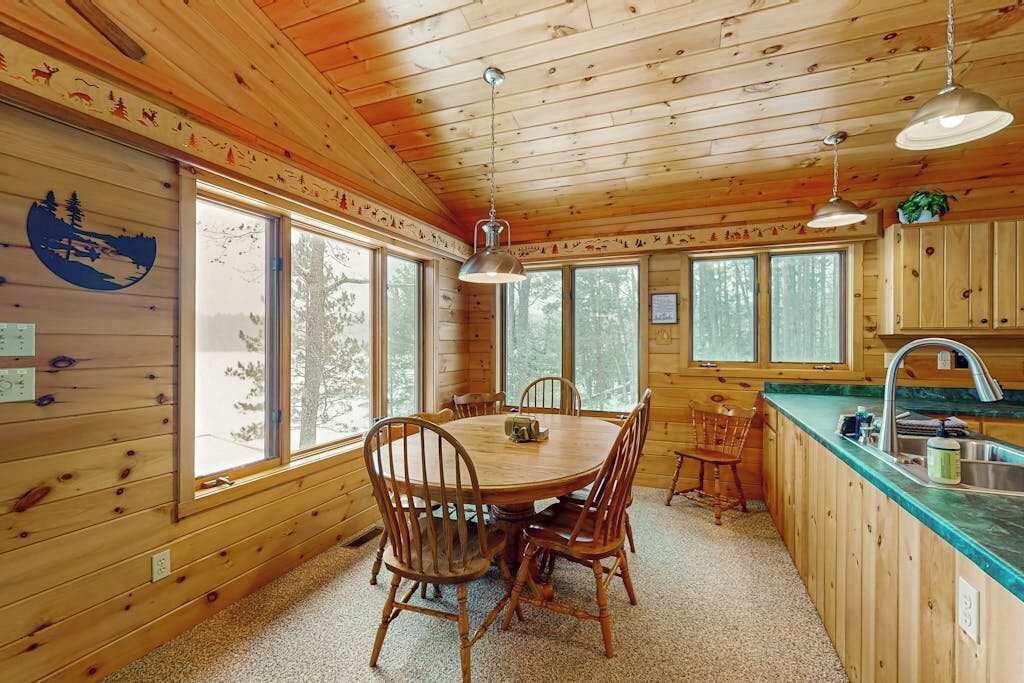 Interior de una cabaña de madera con una mesa de comedor, sillas, una zona de cocina y ventanas amplias.