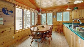 Interior de una cabaña de madera con una mesa de comedor, sillas, una zona de cocina y ventanas amplias.