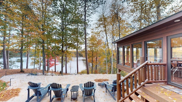 Una terraza con sillas Adirondack y un banco de madera con vista a un lago, rodeado de árboles con follaje otoñal.