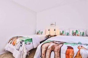 A room with two beds, each with a forest-themed bedspread. There is a small table with two wall-mounted lamps between the beds.